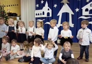 Dzieci 4-letnie śpiewają pastorałkę i grają na janczarach. Za nimi dekoracja: szopka i gwiazda betlejemska, kontur kościoła, domu.